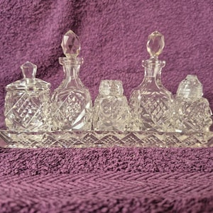 May include: A clear glass vanity set consisting of a rectangular tray, two decanters with faceted stoppers, a lidded jar, and two smaller containers. Each piece features a pressed diamond pattern across the surface, reflecting light through the glass.