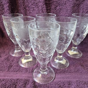 May include: A set of six clear glass goblets featuring a textured, embossed grape and vine pattern on the exterior. Each goblet has a sturdy stem and a circular base, with a consistent design across all pieces, showcasing a detailed fruit motif.