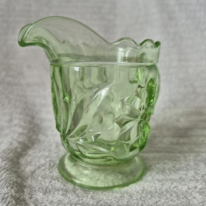 May include: A light green glass creamer with a decorative floral pattern. The creamer has a curved spout and a scalloped rim. The base is round and the glass is transparent. The creamer is approximately 13 cm tall.