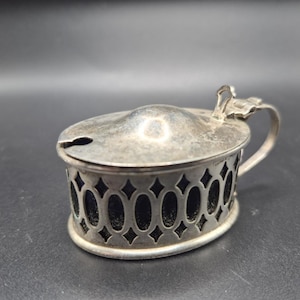 May include: An oval silver-toned metal mustard pot featuring a hinged lid with a small notch for a spoon and a side handle. The body of the container has a decorative openwork lattice pattern with dark blue glass visible through the cutouts.