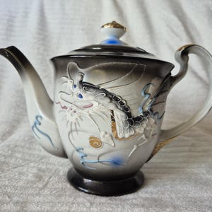 May include: A ceramic teapot featuring a dragon motif. The teapot has a white base with a black and grey gradient towards the top. The dragon is detailed in white, black, and gold, with blue highlights. The handle and spout are white with gold trim.