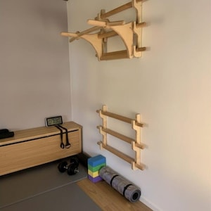 Könnte beinhalten: Holz-Trainingsgeräte an einer weißen Wand. Die Ausrüstung umfasst eine Klimmzugstange und horizontale Stangen. Eine Bank, Hanteln, eine Yogamatte und Schaumstoffblöcke sind ebenfalls zu sehen, was auf ein Home-Gym-Setup hindeutet.