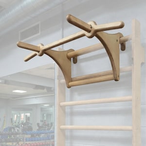 Könnte beinhalten: Holz-Klimmzugstange und Klettergerüst an der Wand befestigt. Die Klimmzugstange ist hellbraun und hat ein einzigartiges Design. Das Klettergerüst hat mehrere horizontale Holzsprossen. Der Hintergrund zeigt eine Turnhalle.