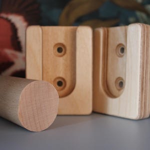 Könnte beinhalten: Drei Holzstücke für ein DIY-Projekt. Zwei Stücke haben einen U-förmigen Ausschnitt und drei Löcher. Das dritte Stück ist ein runder Dübel.