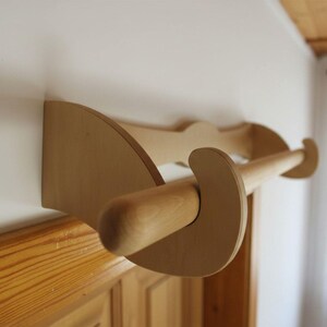 Könnte beinhalten: Ein hellfarbiger Handtuchhalter aus Holz, der an einer weißen Wand befestigt ist. Der Halter hat ein geschwungenes Design mit einer zylindrischen Stange zum Aufhängen von Handtüchern. Die Holzmaserung ist sichtbar, und der Halter ist mit einer flachen, abgerundeten Halterung an der Wand befestigt.