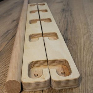 Może przedstawiać: Zestaw drewnianej deski do balansowania, składający się z długiego, cylindrycznego kołka i dwóch pasujących, jasnych drewnianych desek. Każda deska ma wycięte uchwyty i zakrzywioną podstawę, przeznaczoną do ćwiczeń równowagi.