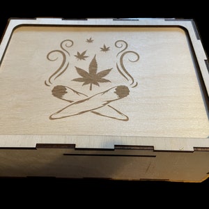 May include: A wooden box with a hinged lid. The lid is decorated with a laser-engraved design of two crossed marijuana joints and a marijuana leaf with smoke coming from the joints.