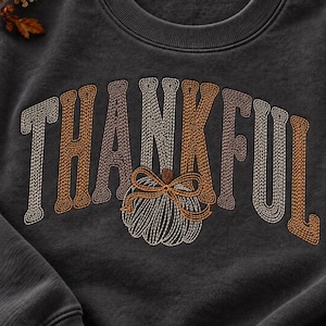 Może przedstawiać: Ciemnoszara bluza z okrągłym dekoltem z wyhaftowanym napisem THANKFUL przy użyciu nici w kolorze kremowym, beżowym i brązowym. Litera K została zastąpiona wyhaftowaną grafiką dyni z małą kokardką w pasujących, neutralnych odcieniach ziemi.