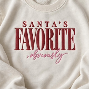 Puede incluir: Una sudadera de cuello redondo color crema con texto bordado en la parte delantera. Las palabras SANTA'S FAVORITE están cosidas en letras mayúsculas rojas en negrita, con la palabra obviously cosida debajo en una fuente cursiva rosa.