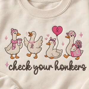 Puede incluir: Una sudadera de cuello redondo color crema con un diseño bordado de cuatro patos con lazos rosas de concienciación sobre el cáncer de mama. El texto "check your honkers" aparece debajo en letra cursiva marrón oscuro.