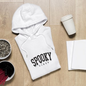 May include: White hooded sweatshirt with the text "Spooky Vibes" printed on the front. The sweatshirt is laid flat on a wooden surface with a cup, two notebooks, and two bowls of sunflower seeds.