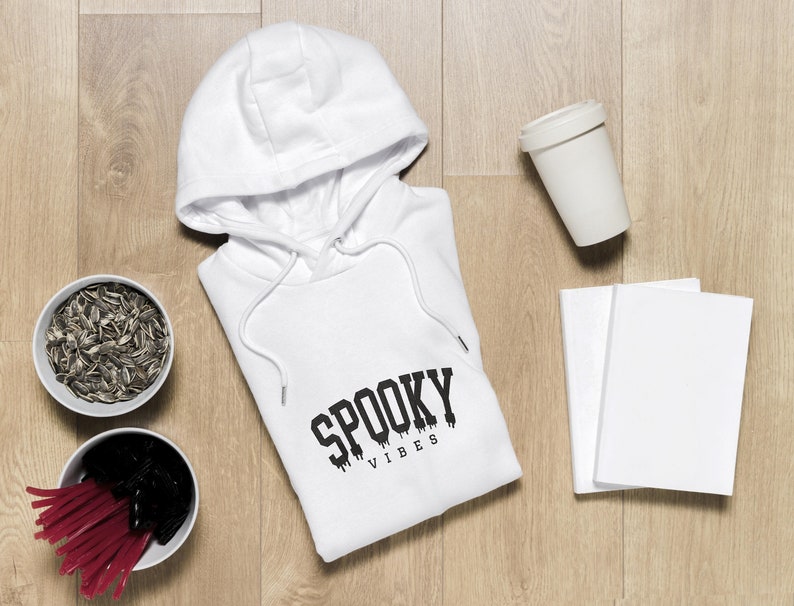 May include: White hooded sweatshirt with the text "Spooky Vibes" printed on the front. The sweatshirt is laid flat on a wooden surface with a cup, two notebooks, and two bowls of sunflower seeds.