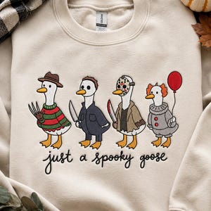 May include: A beige crewneck sweatshirt featuring an embroidered design of four geese dressed as horror movie characters. Below the geese, the text reads "just a spooky goose" in black cursive script. The geese wear costumes including a striped sweater with a hat, a dark jacket with a knife, a hockey mask with a jacket, and a clown outfit with a red balloon.