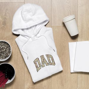 May include: White hoodie with the word "DAD" in a camouflage pattern. The hoodie is on a wooden surface, with two bowls of snacks, a white cup, and two blank notebooks.