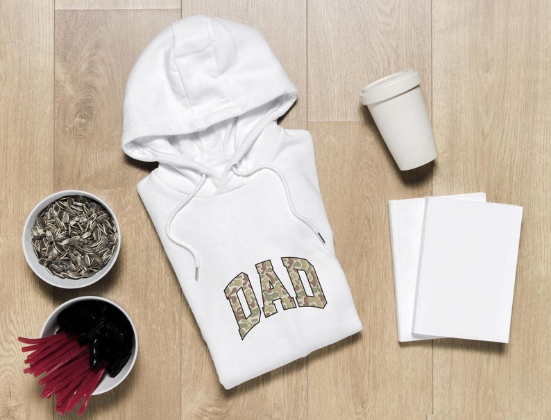 May include: White hoodie with the word "DAD" in a camouflage pattern. The hoodie is on a wooden surface, with two bowls of snacks, a white cup, and two blank notebooks.