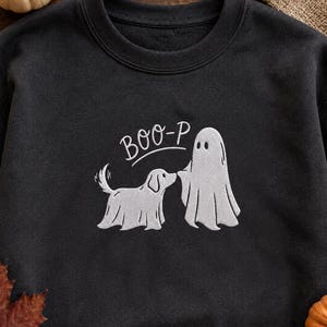 May include: A black crewneck sweatshirt featuring white embroidery of a ghost standing next to a dog wearing a ghost sheet. Above the figures, the text BOO-P is embroidered in a white cursive font, creating a playful Halloween-themed design.