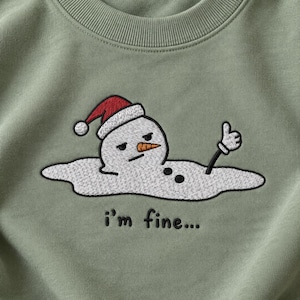 May include: A sage green crewneck sweatshirt featuring an embroidered graphic of a melting snowman wearing a red Santa hat. The snowman has a carrot nose, two black buttons, and one arm raised in a thumbs-up gesture. The text i'm fine... is embroidered below.