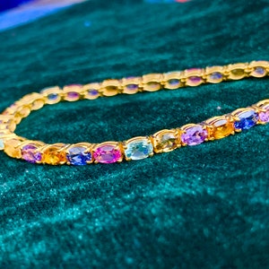 Puede incluir: Una pulsera dorada con una serie de piedras preciosas de corte ovalado y coloridas. Las piedras muestran un espectro de colores, incluyendo azul, morado, amarillo y rosa. La pulsera está sobre un fondo verde oscuro.