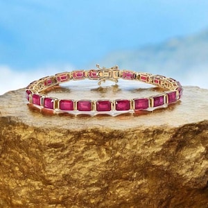 Könnte beinhalten: Ein goldenes Armband mit einer Reihe von roten quadratischen Edelsteinen. Das Armband liegt auf einer goldfarbenen Oberfläche.