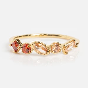 Könnte beinhalten: Ein goldfarbener Ring mit einer Reihe farbiger Edelsteine. Der Ring zeigt quadratische, runde und ovale Steine in Orange- und Pfirsichtönen. Das Band hat ein strukturiertes Aussehen.