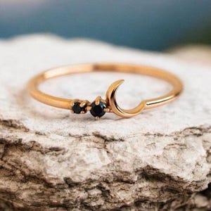 Könnte beinhalten: Ein zarter Goldring mit einem Halbmond-Design und zwei kleinen schwarzen Edelsteinen. Der Ring liegt auf einer rauen, strukturierten Oberfläche.