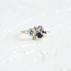 Könnte beinhalten: Ein goldener Ring mit einem Cluster aus blauen, grünen und violetten Edelsteinen. Der Ring liegt auf einem weißen Hintergrund.