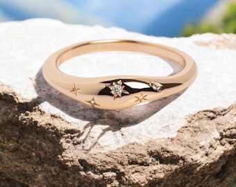 May include: A gold-colored ring with a smooth, curved design. The ring is adorned with small star-shaped embellishments and a central, larger starburst design with a clear stone. The ring is displayed on a textured, light brown surface.