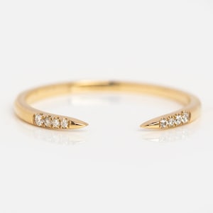 Könnte beinhalten: Ein goldfarbener offener Ring mit minimalistischem Design. Der Ring hat kleine, funkelnde Edelsteine, die entlang des Bandes in der Nähe der spitzen Enden eingesetzt sind. Der Ring wird auf einer weißen Oberfläche präsentiert.