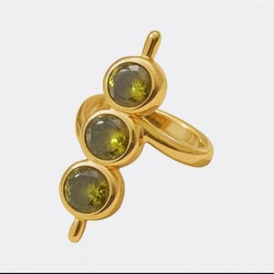 Puede incluir: Un anillo dorado con tres piedras preciosas redondas de color verde oliva. El anillo tiene un diseño único con las piedras dispuestas verticalmente, cada una engastada en un bisel dorado. La banda del anillo es lisa y pulida, creando un aspecto moderno y elegante.