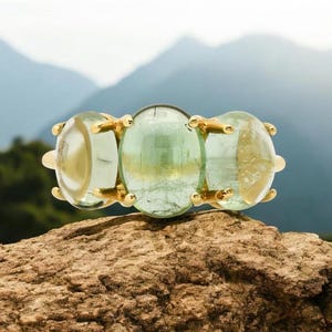 May include: A gold ring featuring three oval-shaped green gemstones. The ring is resting on a rock with a blurred background of mountains.