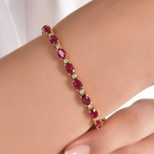 Könnte beinhalten: Ein goldenes Armband mit oval geschliffenen roten Edelsteinen und kleinen, quadratisch geschliffenen weißen Edelsteinen. Das Armband wird an einem Handgelenk getragen, wodurch das Design und der Farbkontrast hervorgehoben werden. Schmuck.