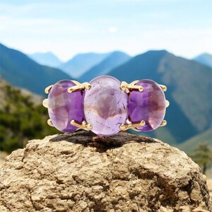 Puede incluir: Un anillo de oro con tres grandes piedras de amatista de forma ovalada. El anillo está colocado sobre un fondo de montañas y cielo azul.