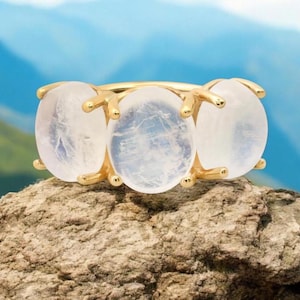 Puede incluir: Un anillo de oro con tres piedras de luna ovaladas. El anillo está colocado sobre una roca con un fondo azul y verde.