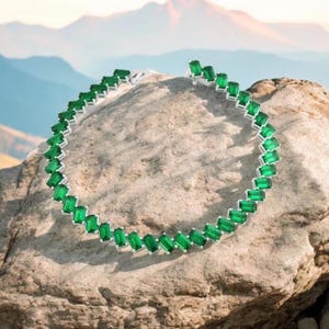 Puede incluir: Un collar de plata con piedras rectangulares de color verde esmeralda. El collar está sobre una roca con un fondo de montaña.