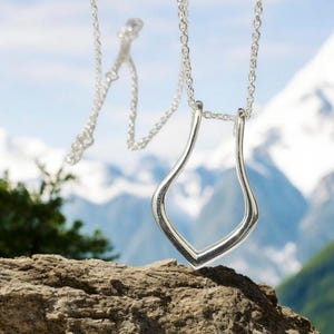 Puede incluir: Un collar de plata con un colgante único y abstracto en forma de lágrima. El colgante está colgando sobre un fondo de una cordillera.
