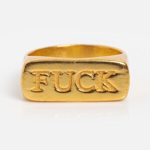 Op de afbeelding: Een goudkleurige ring met het woord "FUCK" in reliëf op de bovenkant. De ring heeft een rechthoekige voorkant en een gladde, gepolijste afwerking. De letters zijn verhoogd en in een vetgedrukte, schreefloze lettertype.