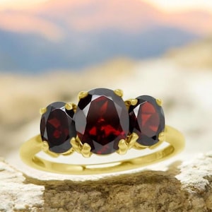 May include: A gold ring featuring three oval-cut, deep red gemstones. The gemstones are set in a gold band, with the center stone slightly larger than the two flanking stones. The ring is displayed on a textured surface.