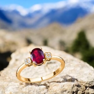 Könnte beinhalten: Ein goldener Ring mit einem großen ovalen Rubin-Edelstein und zwei kleineren Diamant-Akzenten.
