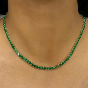 Könnte beinhalten: Eine goldene Halskette mit einer Reihe leuchtend grüner Edelsteine. Die Halskette, die um den Hals getragen wird, zeigt smaragdähnliche Steine, die in einer Goldkette gefasst sind. Die Edelsteine sind eng aneinander gesetzt und bilden eine durchgehende Farblinie.