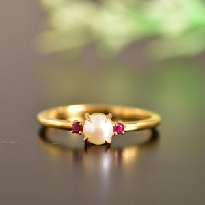 Könnte beinhalten: Ein goldener Ring mit einer weißen Perle und zwei kleinen roten Edelsteinen.