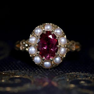 Könnte beinhalten: Ein goldener Ring mit einem großen ovalen rosafarbenen Edelstein, der von einem Heiligenschein aus weißen Perlen umgeben ist.