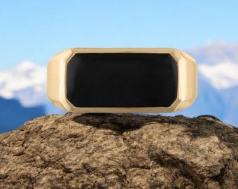 May include: A gold ring with a black rectangular stone set in the center. The ring is sitting on a rock.