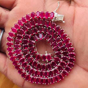 May include: A necklace featuring a series of oval-cut, deep red gemstones set in a silver-toned metal. The gemstones are closely arranged, creating a continuous, vibrant display. The necklace is secured with a clasp.
