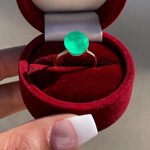 Könnte beinhalten: Ein goldener Ring mit einem großen, runden, leuchtend grünen Edelstein, präsentiert in einer offenen roten Samt-Ringbox. Der Ring wird von einer Person mit einer manikürten Hand gehalten.