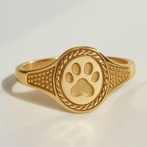 Könnte beinhalten: Ein goldfarbener Ring mit einem Pfotenabdruck-Design. Der Ring hat eine kreisförmige Mitte mit einem detaillierten Pfotenabdruck, umgeben von einem seilartigen Rand. Das Band hat strukturierte Details an den Seiten, was einen klassischen und eleganten Look erzeugt.