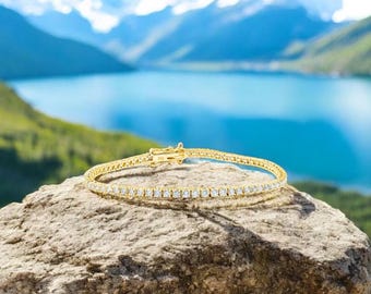 May include: A gold tennis bracelet with a row of small, clear diamonds. The bracelet is on a rock with a lake and mountains in the background.