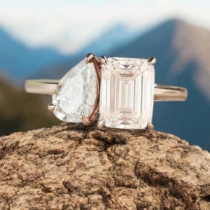Könnte beinhalten: Ein Ring mit einem birnenförmigen Diamanten und einem Smaragdschliff-Diamanten, gefasst in einer goldenen Fassung. Der Ring liegt auf einem Stück grobem Holz.