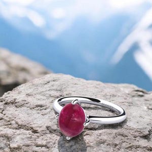 Puede incluir: Un anillo de plata con una piedra ovalada de color rosa brillante engastada en garras. El anillo está sobre una roca gris.
