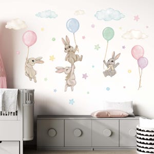 K&ouml;nnte beinhalten: Ein Set aus vier Wandaufklebern in Pastellfarben mit Ballons und Hasen. Die Hasen halten die Ballons fest und fliegen durch die Luft. Es gibt auch Wolken und Sterne im Design.