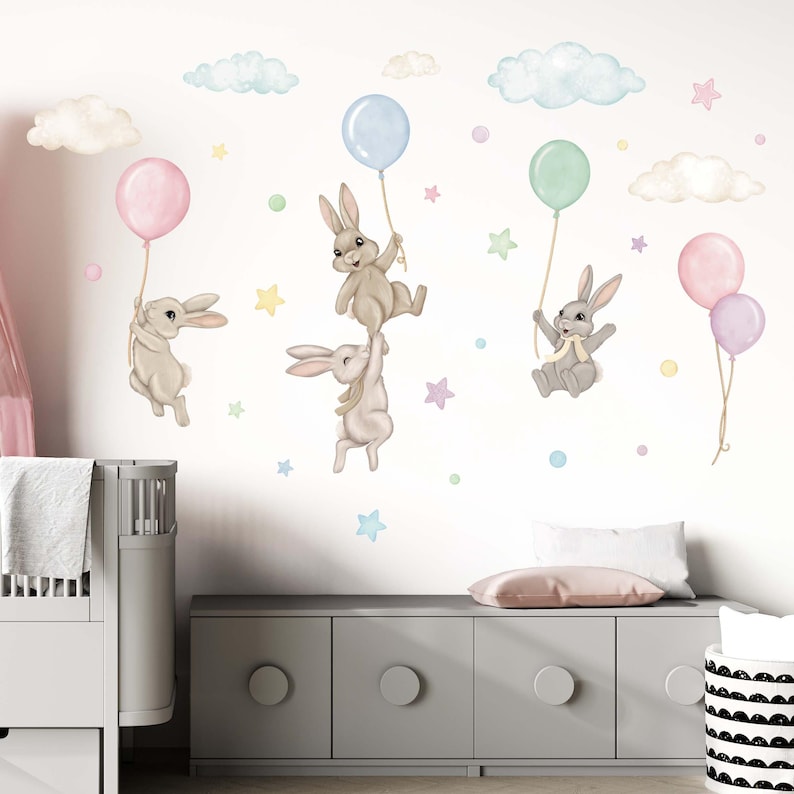 K&ouml;nnte beinhalten: Ein Set aus vier Wandaufklebern in Pastellfarben mit Ballons und Hasen. Die Hasen halten die Ballons fest und fliegen durch die Luft. Es gibt auch Wolken und Sterne im Design.